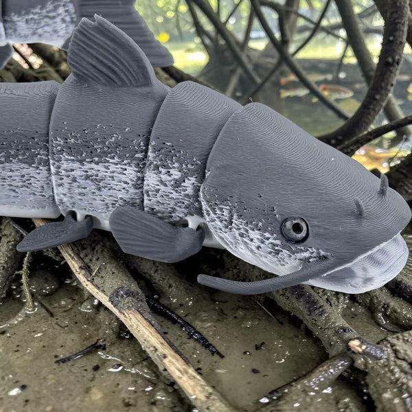 Tidal 3D Printed articulated grey catfish toy with realistic mottled texture and barbels resting on muddy ground.