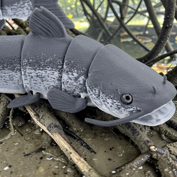 Tidal 3D Printed articulated grey catfish toy with realistic mottled texture and barbels resting on muddy ground.